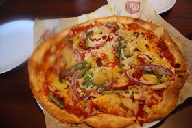 A pizza with Dairy Free Cheese, Tomato Sauce, Chorizo, Grilled Chicken, Bacon, Cauliflower, Asparagus, Pineapple, Red Onion, and Peppers.