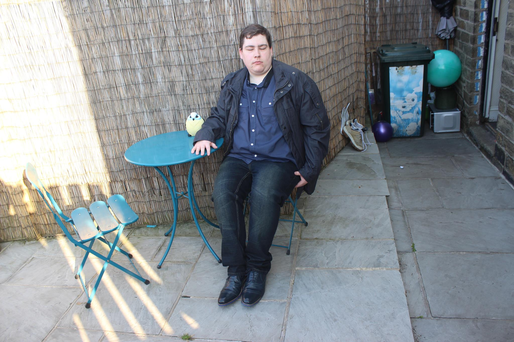 Me sat in a garden chair wearing a jacket, a short sleeved shirt, a pair of jeans, and some shoes.