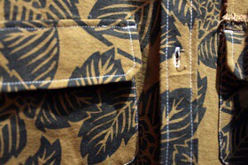 A brown shirt, with green leaf detailing, zoomed in, on a grey background.
